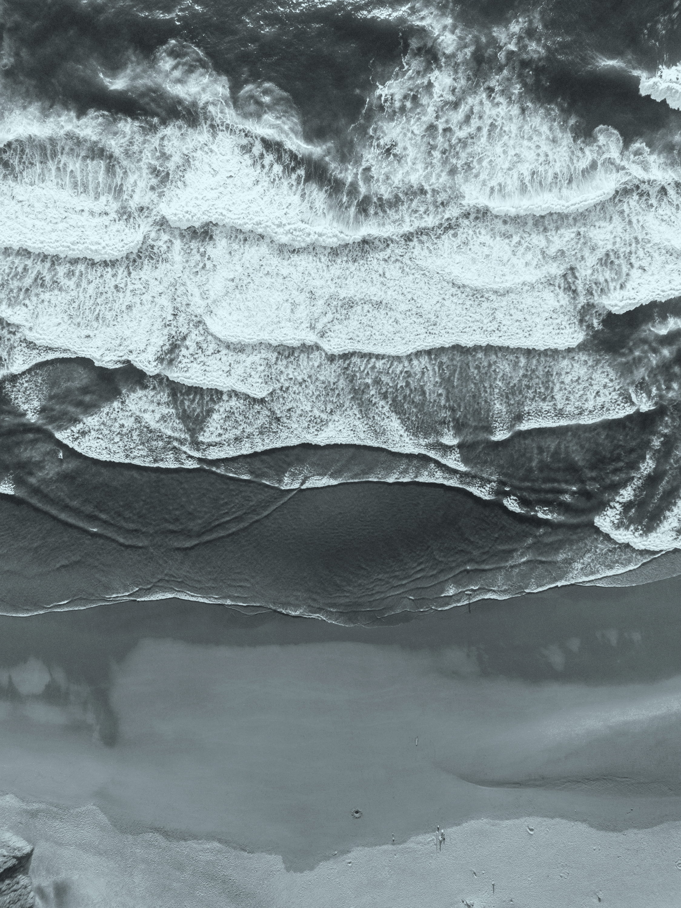 Aerial view of ocean waves crashing onto a sandy beach, captured in black and white.