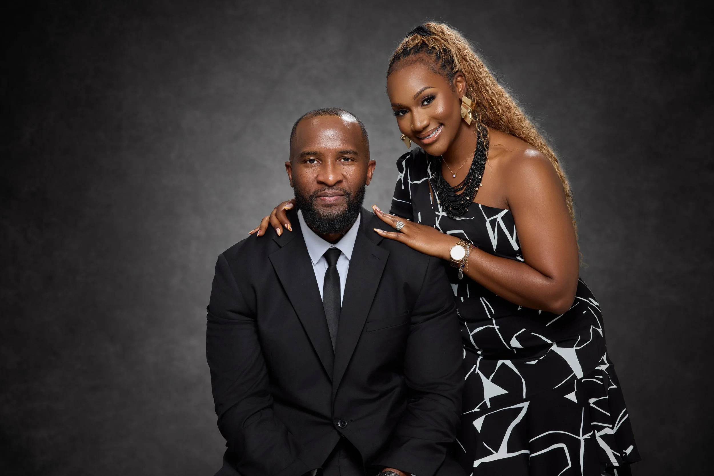 A man in a black suit and tie sitting next to a woman in a black and white patterned dress, both smiling against a dark background.