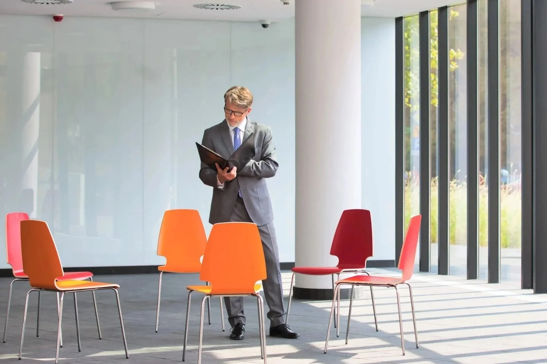 A man in a gray suit and glasses stands in a modern, spacious room with large windows, looking at a folder, surrounded by colorful chairs.