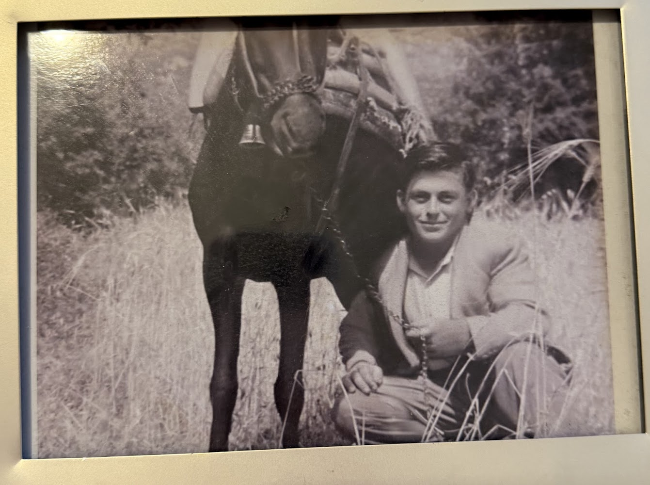 A black and white photo of a young man with short hair, crouching next to a black dog in a grassy outdoor area with trees in the background.