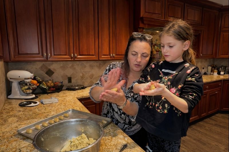 A woman and a young girl baking together in a kitchen. The woman is guiding the girl in shaping dough, with a tray of dough pieces and a mixing bowl on the counter.