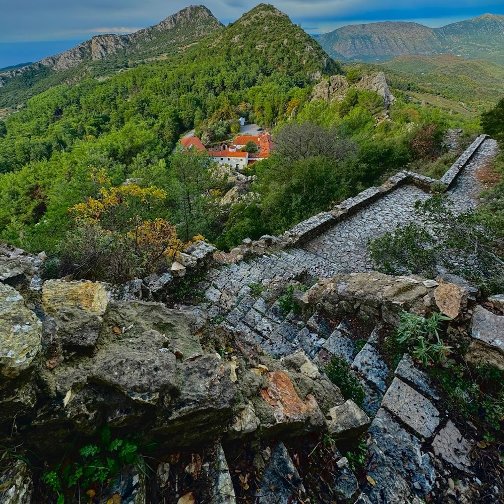 Stone pathway and ruins overlooking lush green forest and distant mountains, with a small village visible among the trees.