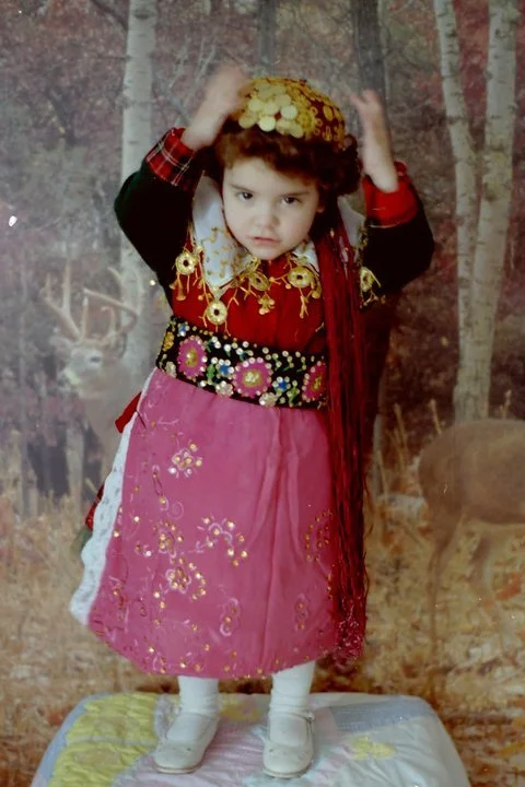 Young girl dressed in colorful traditional costume, standing on a cloth-covered platform, holding a large yellow headdress with coins, in front of a forest backdrop.