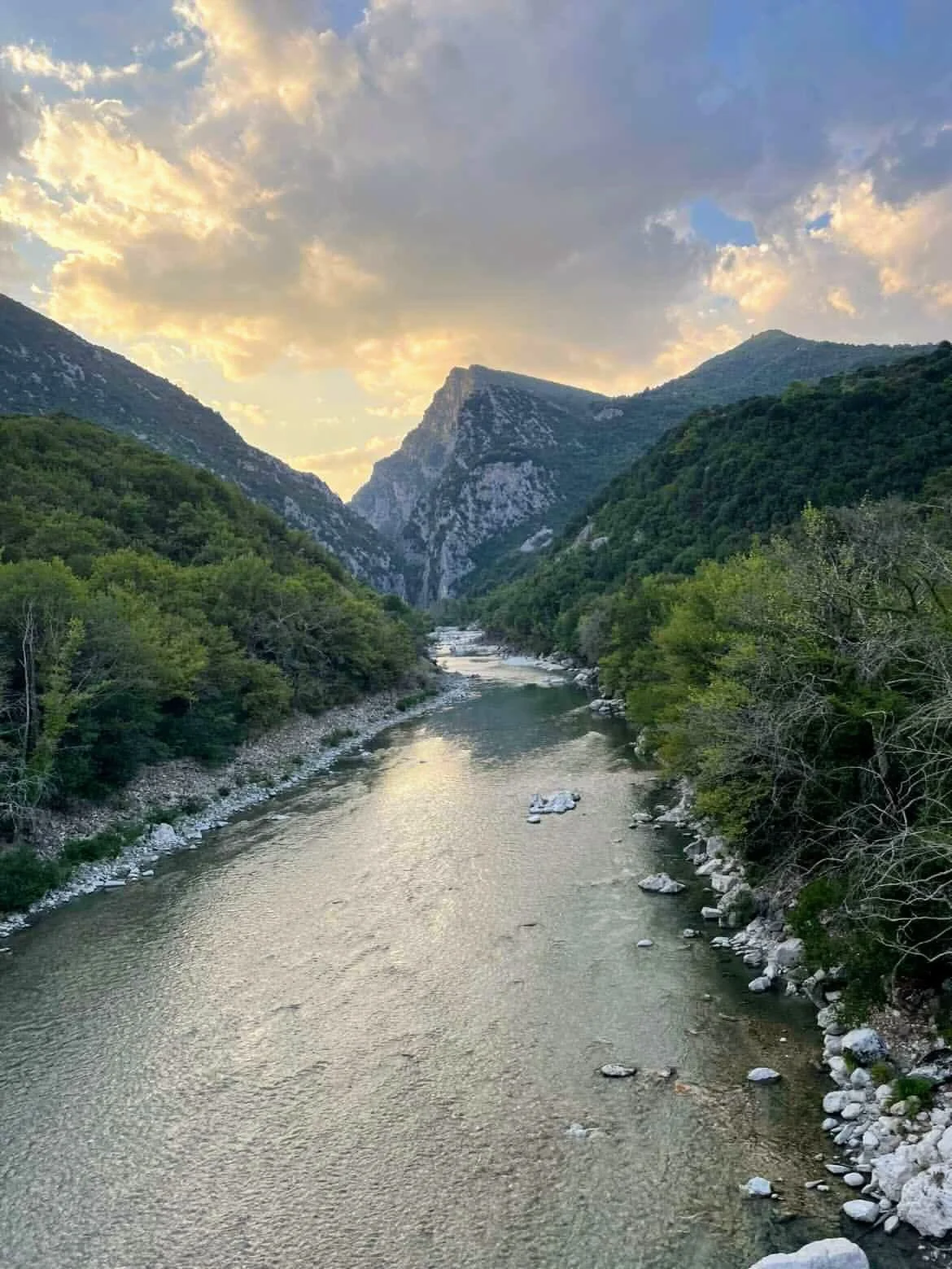 A river flowing between lush green mountains with a partly cloudy sky overhead at sunset.