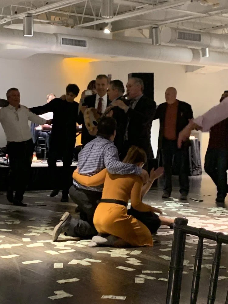 People celebrating at an event, with two individuals kneeling on the floor surrounded by scattered papers, while others dance and watch in the background.