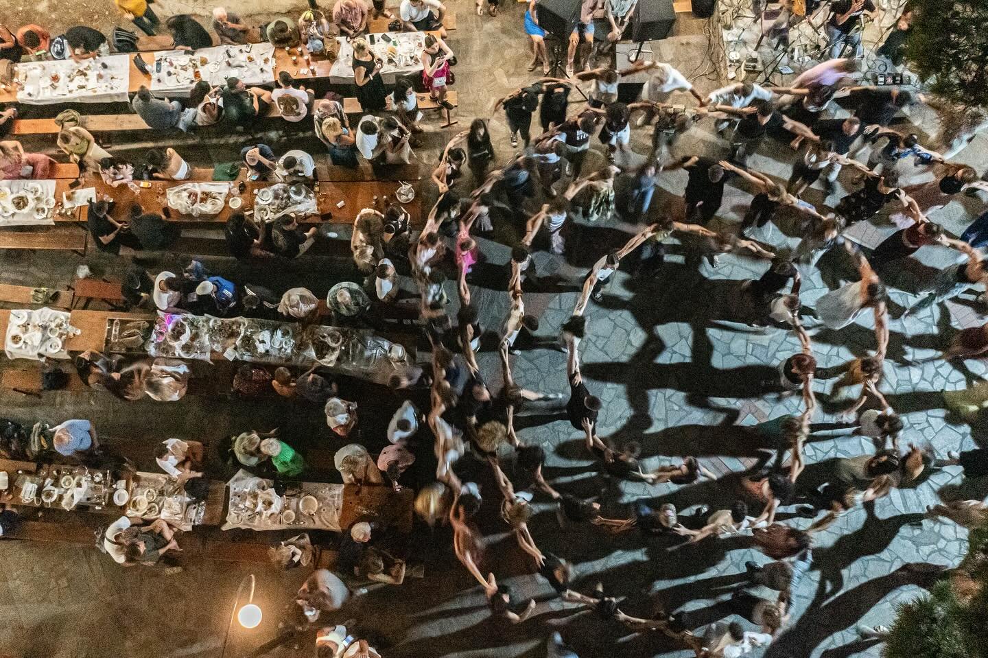 People sitting at tables and standing in lines in an outdoor setting, with a group of individuals forming a human chain and casting long shadows at night.
