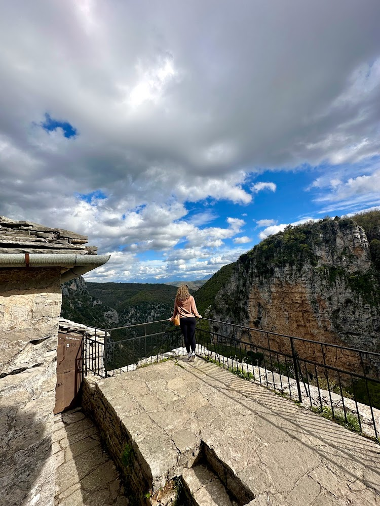 A woman standing on a stone-floored viewing platform with black metal railings, overlooking a deep canyon with cliffs and lush greenery under a partly cloudy sky.