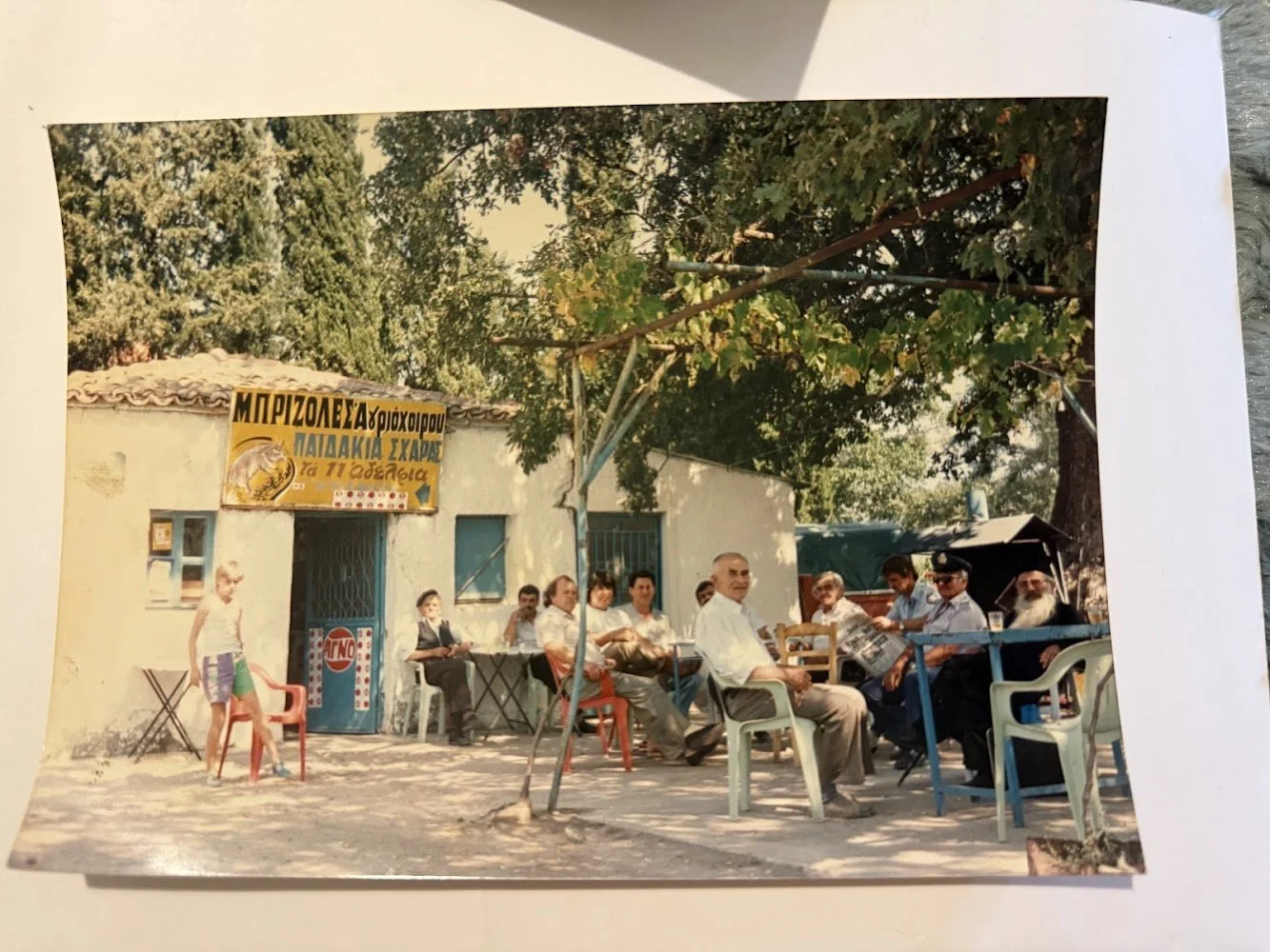 A group of men and a young girl sitting and standing outside a small building with a yellow sign, trees in the background, and outdoor tables and chairs.