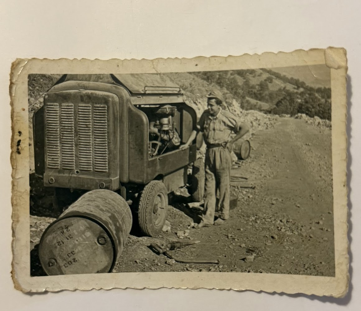 A black and white photo of a man in military uniform standing next to a vintage truck with its hood open, in a rugged outdoor setting with hills in the background. A barrel is lying on its side nearby.