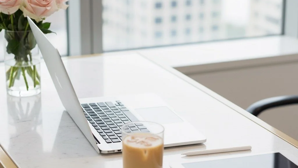 Laptop on a white table with a glass of iced coffee, a pen, and a vase of flowers, in front of a window with a city view.