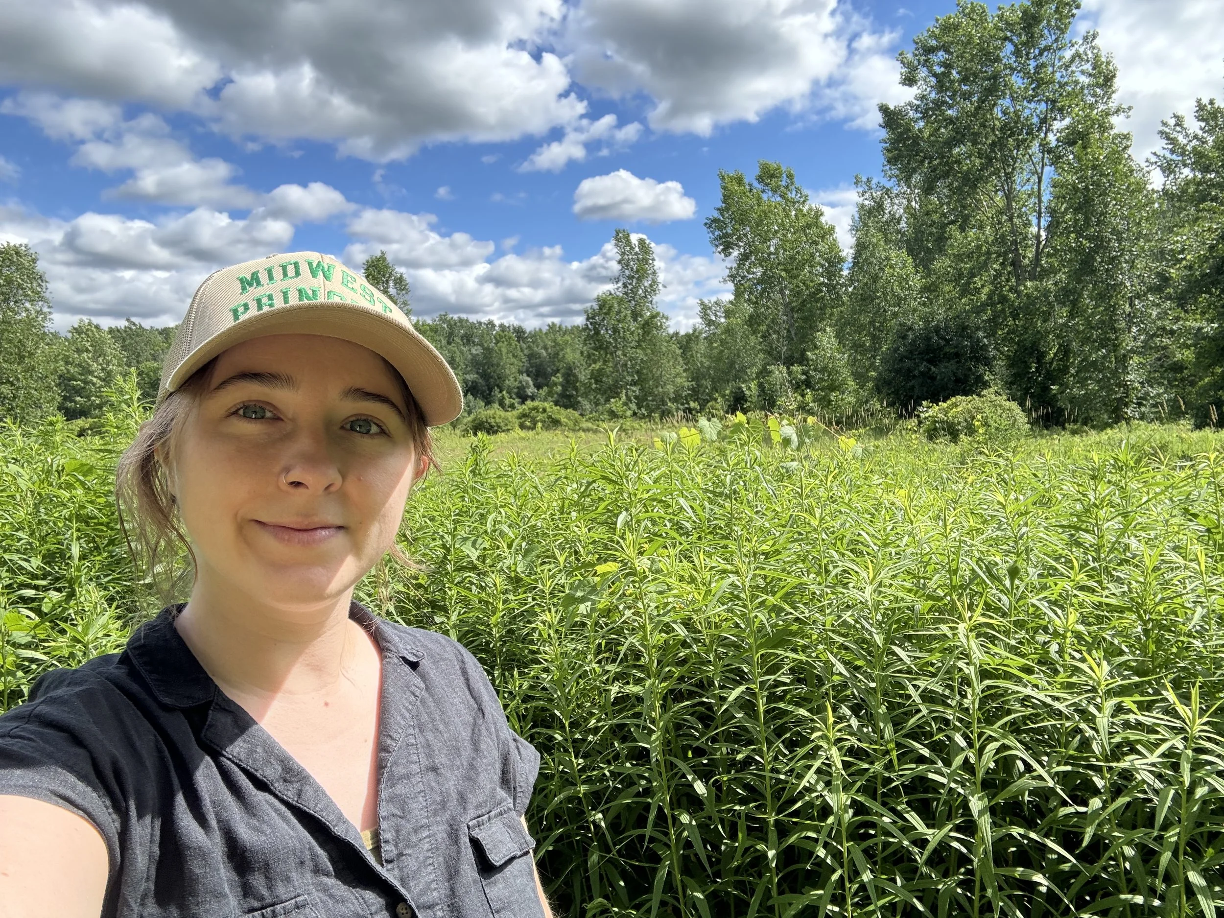 A woman taking a selfie in a green field with lush plants, trees in the background, and a partly cloudy blue sky.