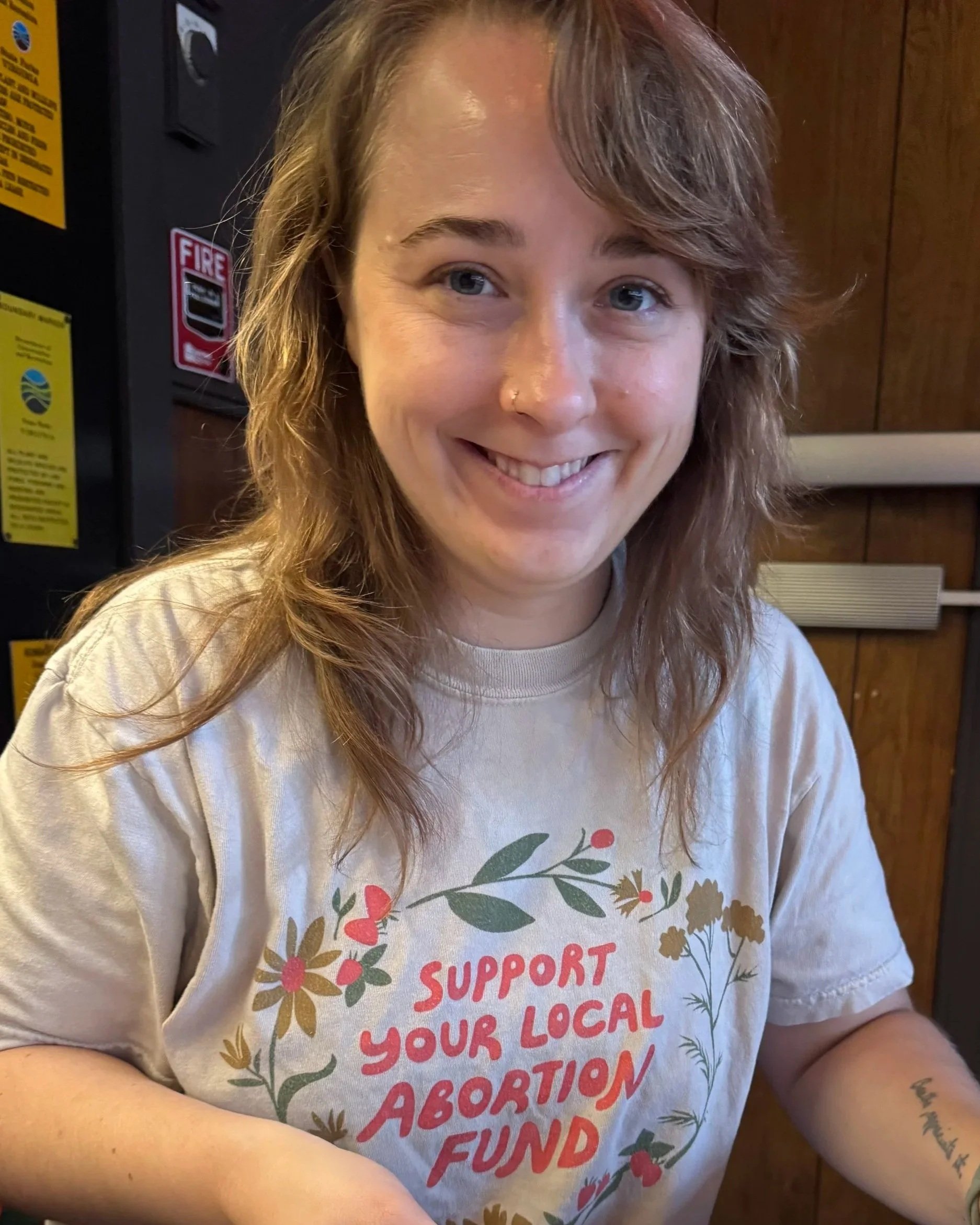 white person long brown wavy hair smiles at camera shirt says support your local abortion fund
