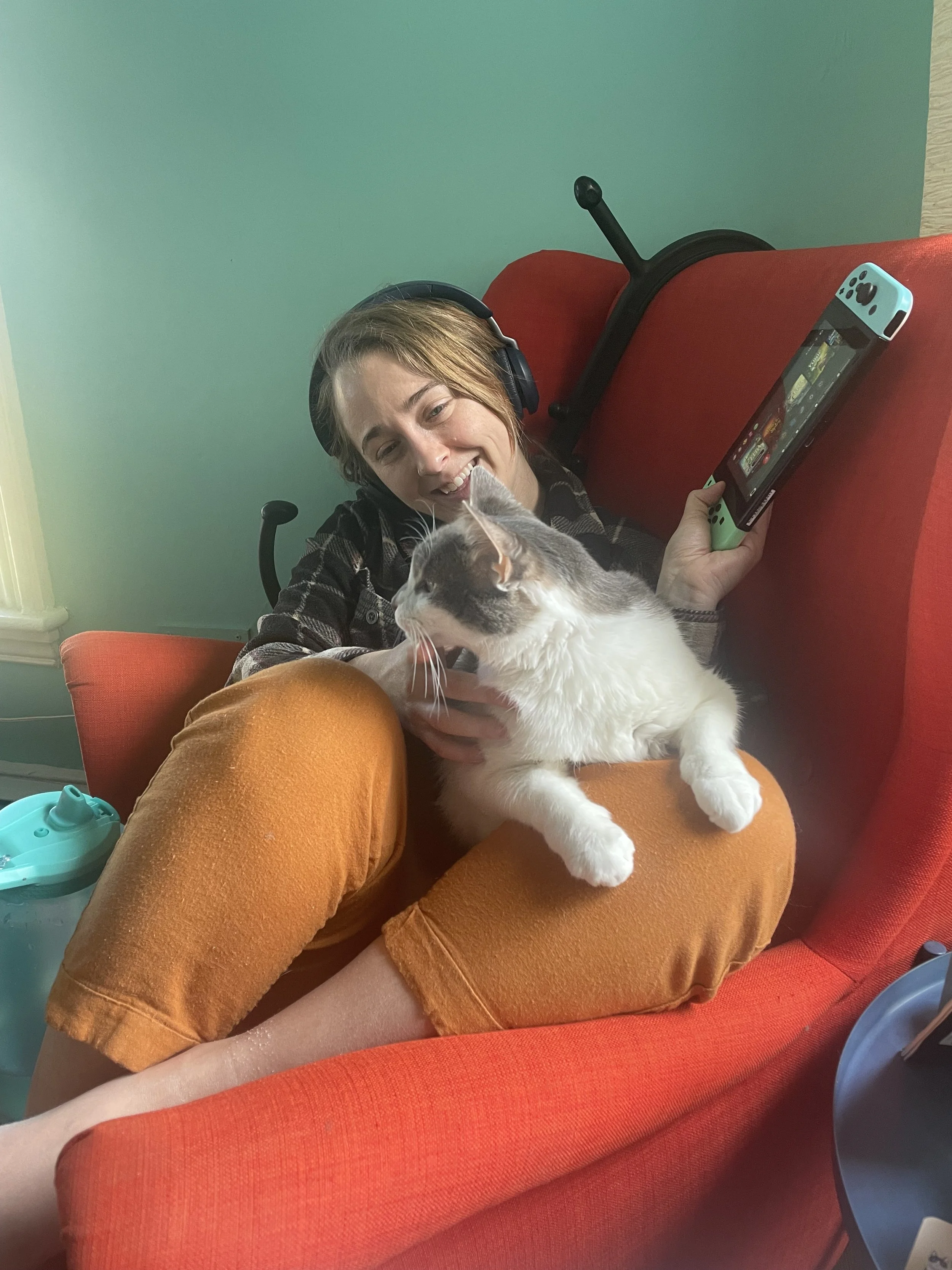 Person sits curled up on a chair switch in hand with grey and white cat sitting on lap