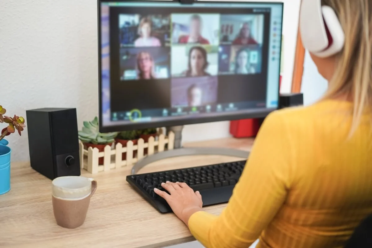 Person in yellow shirt wearing headphones on a video call at a computer desk with potted plants and speakers.