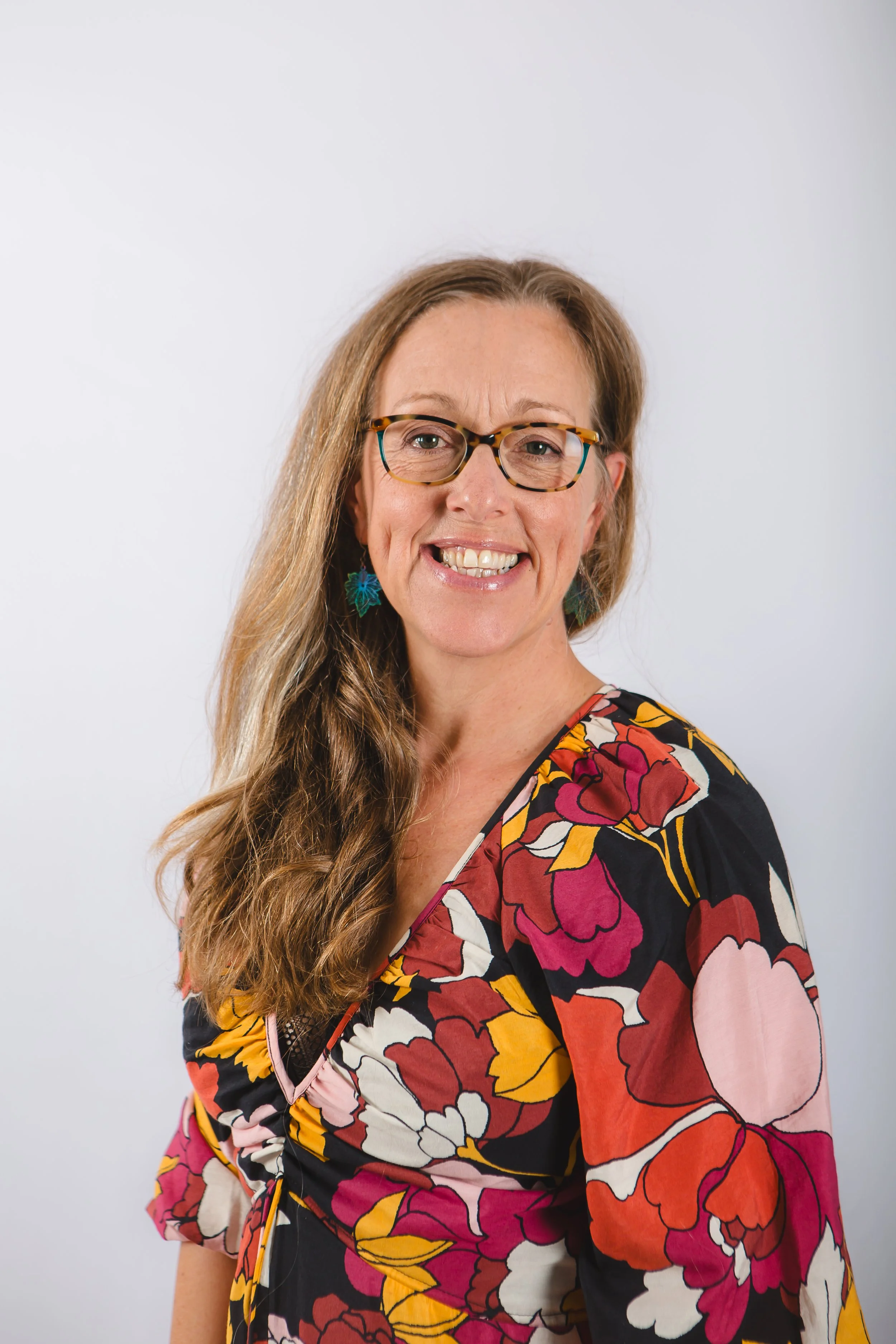 A smiling woman with brown hair, glasses, and colorful floral dress poses against a plain white background.