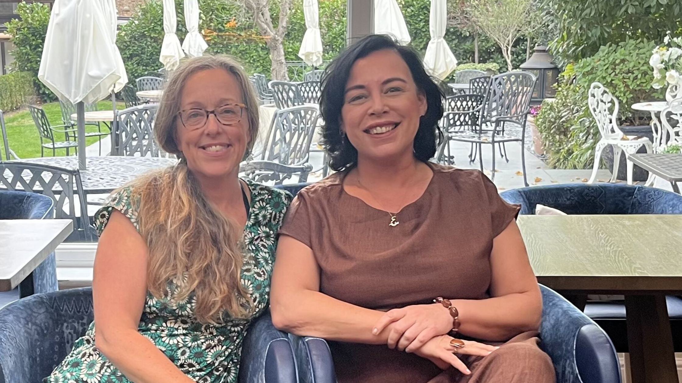 Two women sitting at a table in an outdoor dining area, smiling at the camera. One has long, wavy blonde hair and glasses, and the other has shoulder-length black hair, wearing a brown top with a letter 'L' necklace.