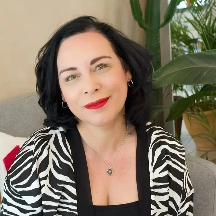 A woman with black hair, wearing red lipstick, silver hoop earrings, and a zebra-print jacket, sitting indoors with green plants in the background.