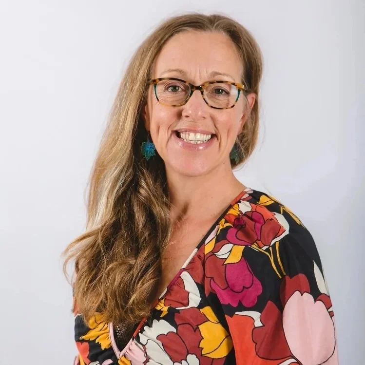 A woman with long wavy hair, glasses, and earrings, smiling and wearing a colorful floral dress against a plain background.