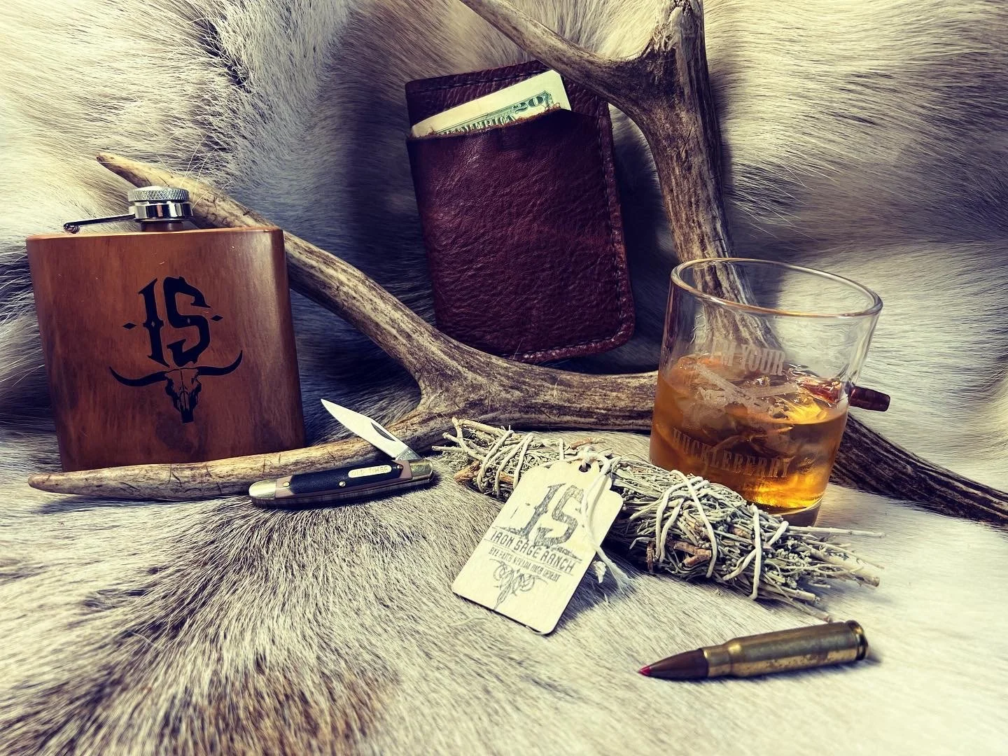A collection of objects with a cowboy or western theme displayed on animal fur background includes a wooden flask with a skull logo, a leather wallet with dollar bill, a glass of whiskey with ice, antler pieces, a knife, a bundle of sage or herbs tie