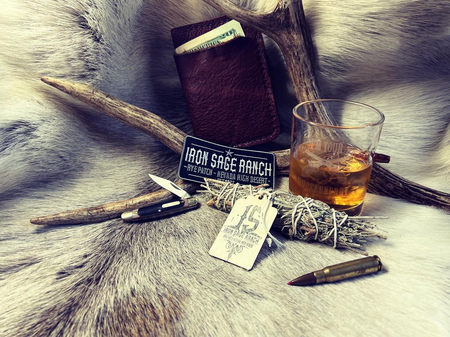 A rustic scene featuring a whiskey glass with ice, a leather wallet, a fixed-blade knife, a small silver bullet, a bundle of sage tied with string, a patch with the text 'Iron Sage Ranch', a tag, and a pair of antlers on animal fur.