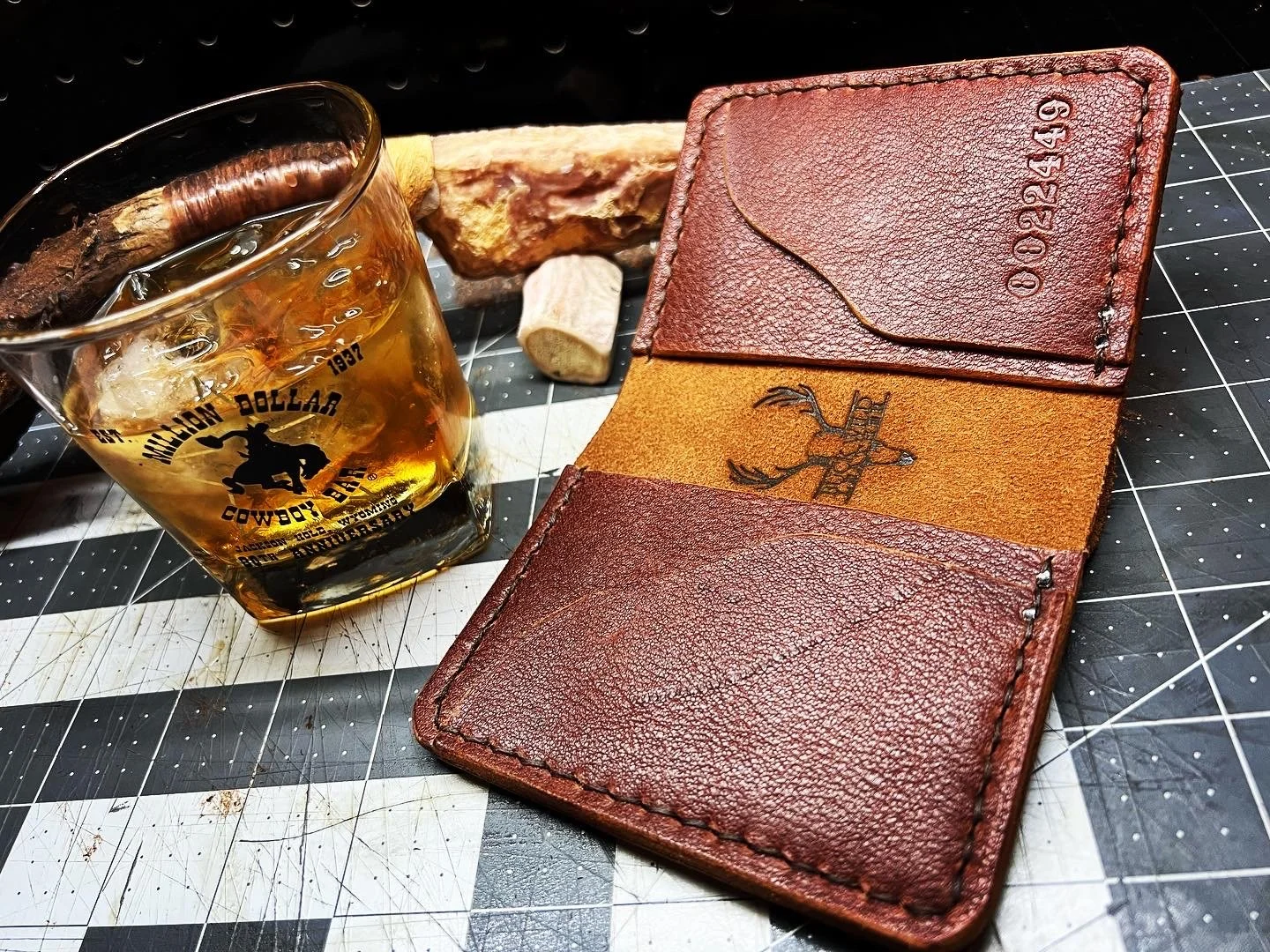 A glass of whiskey with ice, a leather wallet, and a piece of wood and rock on a cutting mat.