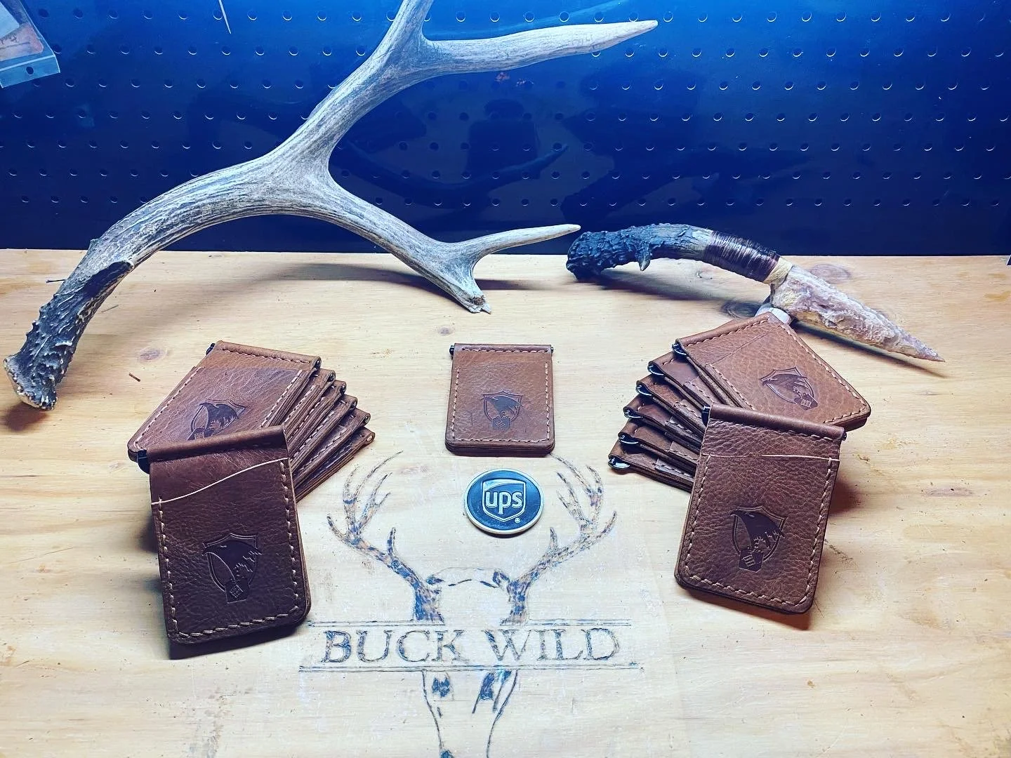Several leather wallet holders with Buck Wild logo, a UPS badge, deer antlers, and a deer skull on a wooden surface.