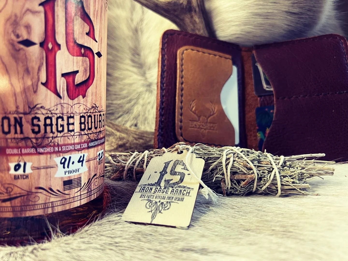 A bottle of bourbon whiskey, a bundle of dried sage tied with a string, a leather wallet, and a flask with a colorful design, all placed on a soft fur surface.