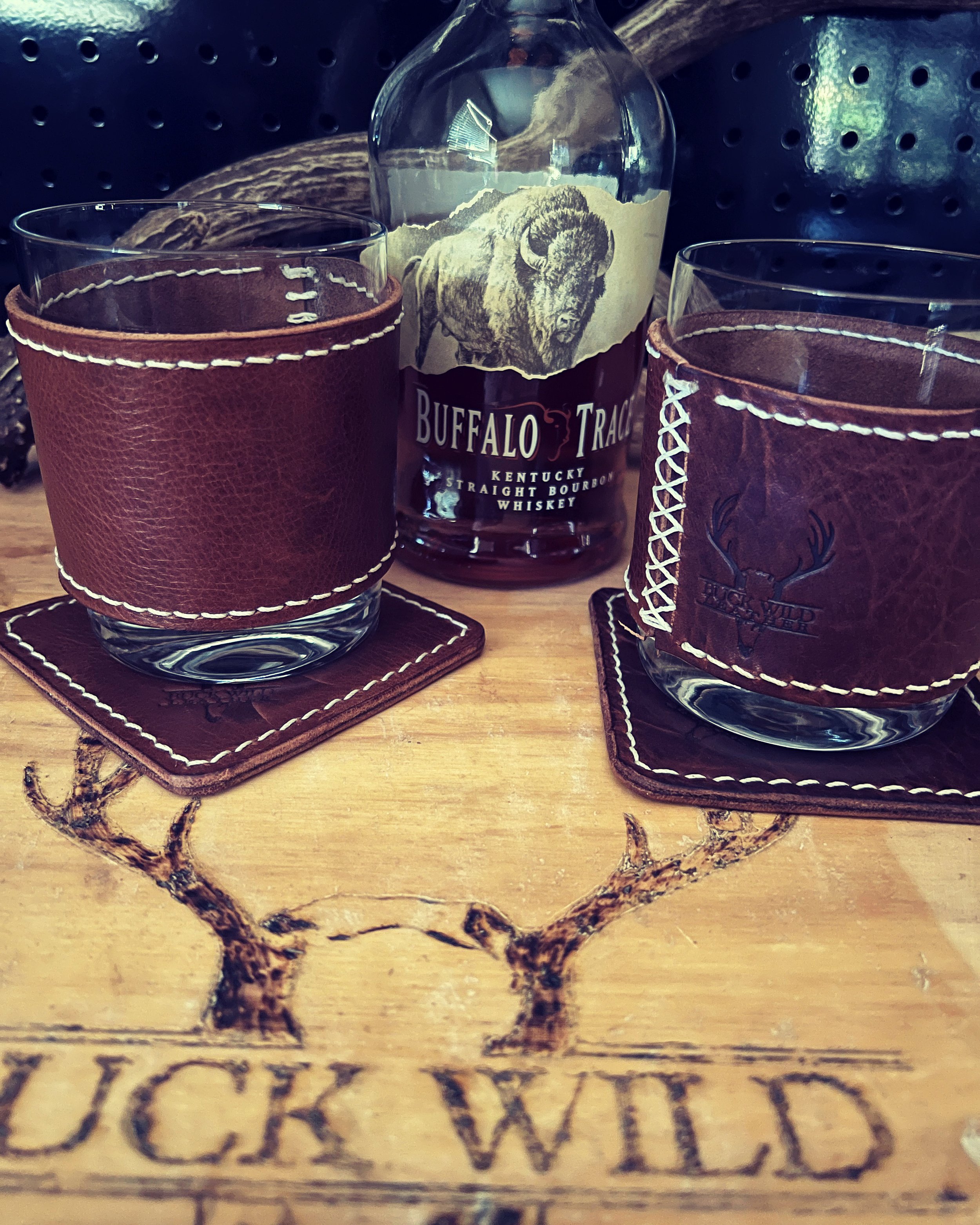 Two whisky glasses with leather holders and a bottle of Buffalo Trace bourbon on a wooden table with Buck Wild engraved, decorated with antler designs.
