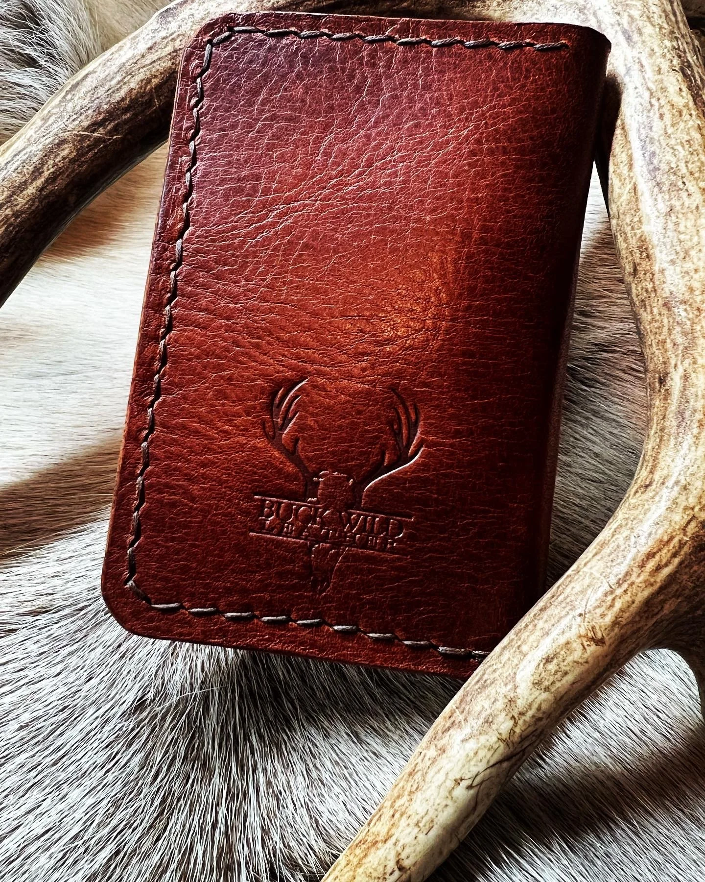 Close-up of a brown leather wallet with an embossed deer head logo and the words 'BISON WILD' on it, resting on a fur surface with a curved horn nearby.
