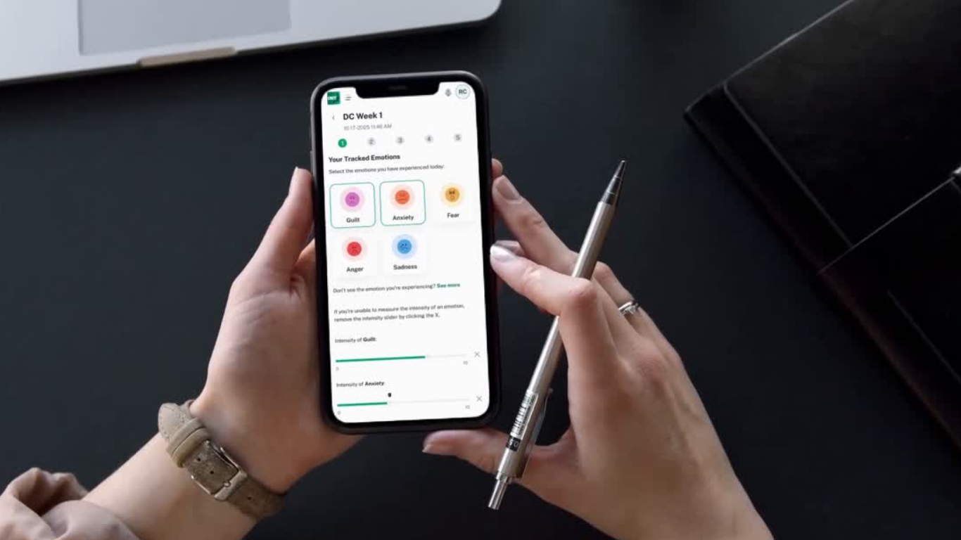 A person holding a smartphone and a pen while looking at an app tracking emotions like guilt, anxiety, fear, anger, and sadness, on a black desk with a laptop and other office items nearby.