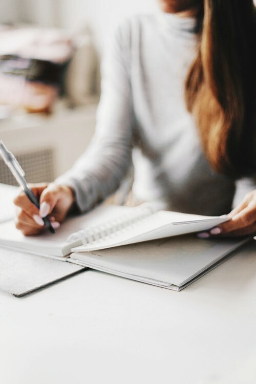 A woman wearing a gray long-sleeve shirt writing in a notebook at a desk.