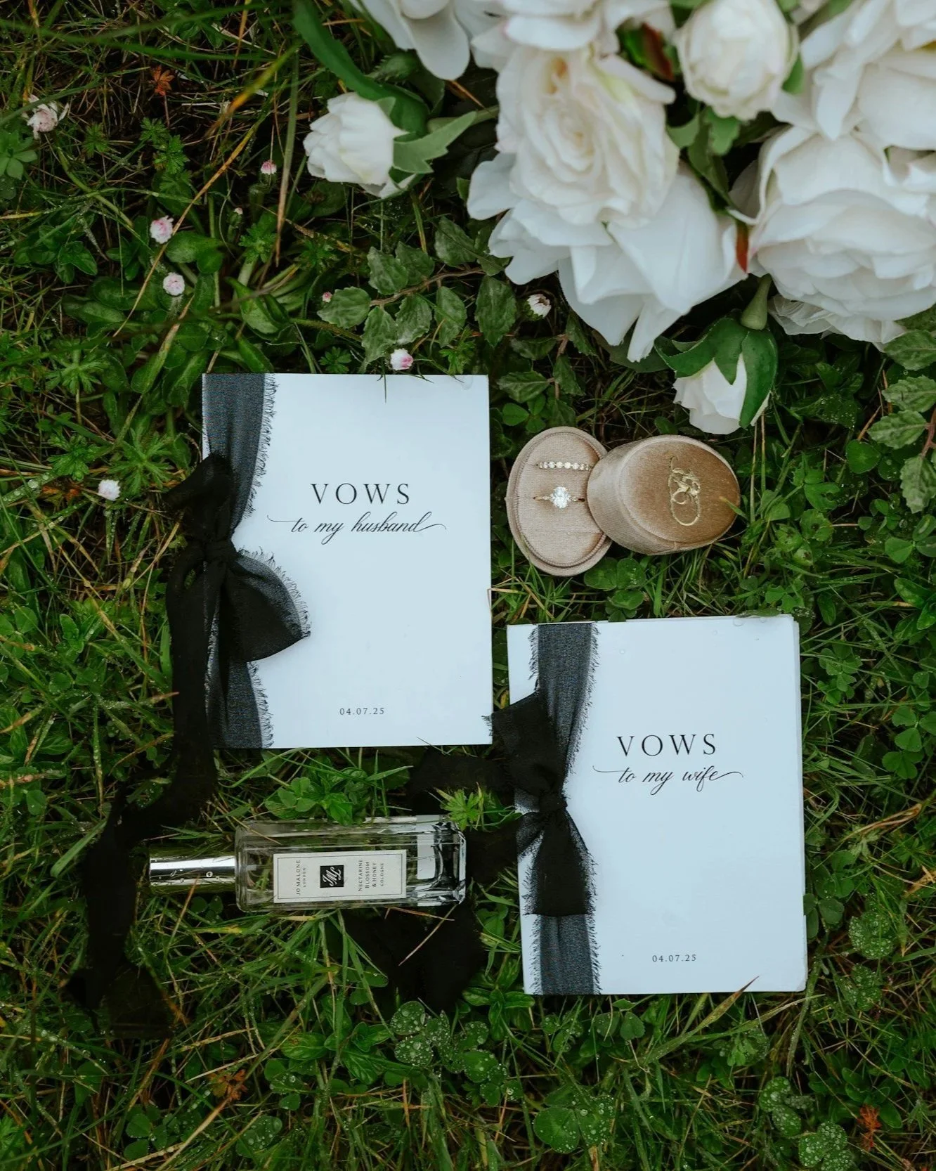 Wedding vows booklets, one labeled "VOWS to my husband" and the other "VOWS to my wife," decorated with black ribbons, surrounded by white flowers, greenery, and a small perfume bottle.
