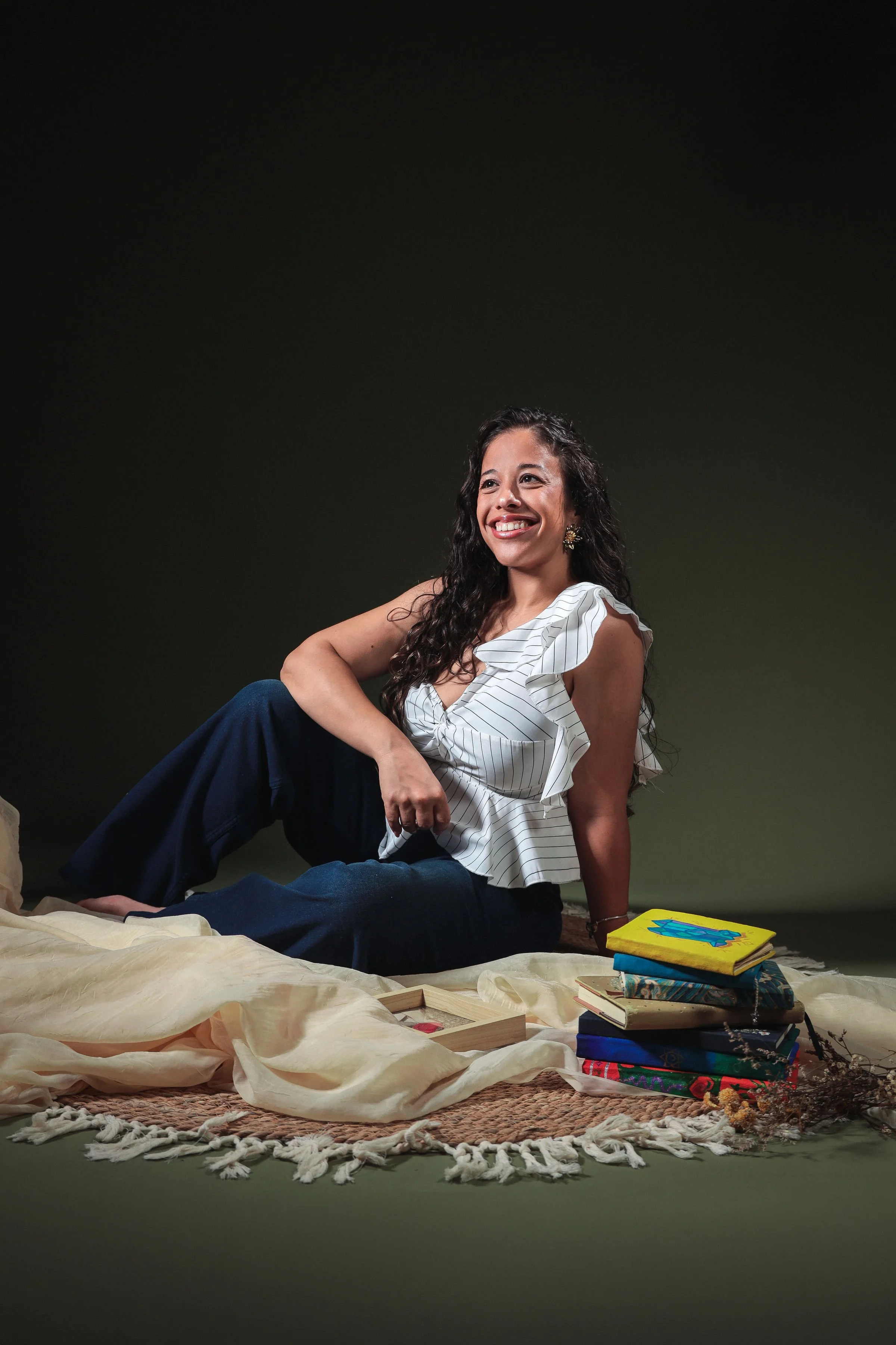 A woman with long curly hair sitting on a carpeted floor against a dark background. She is smiling, looking to her right, wearing a white ruffled top and dark pants. There are colorful books and a box near her on the floor, with fabric and decorative items around.