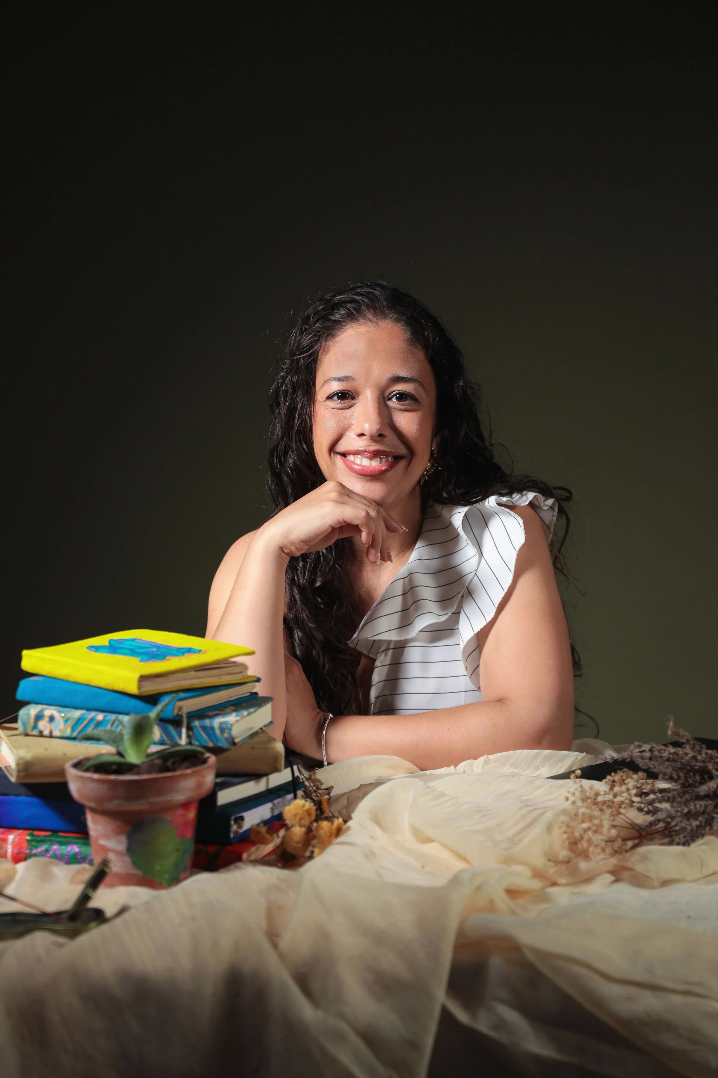 A woman with long, dark, curly hair smiling with her chin resting on her hand, surrounded by a stack of books, plants, and dried flowers on a table, against a dark background.