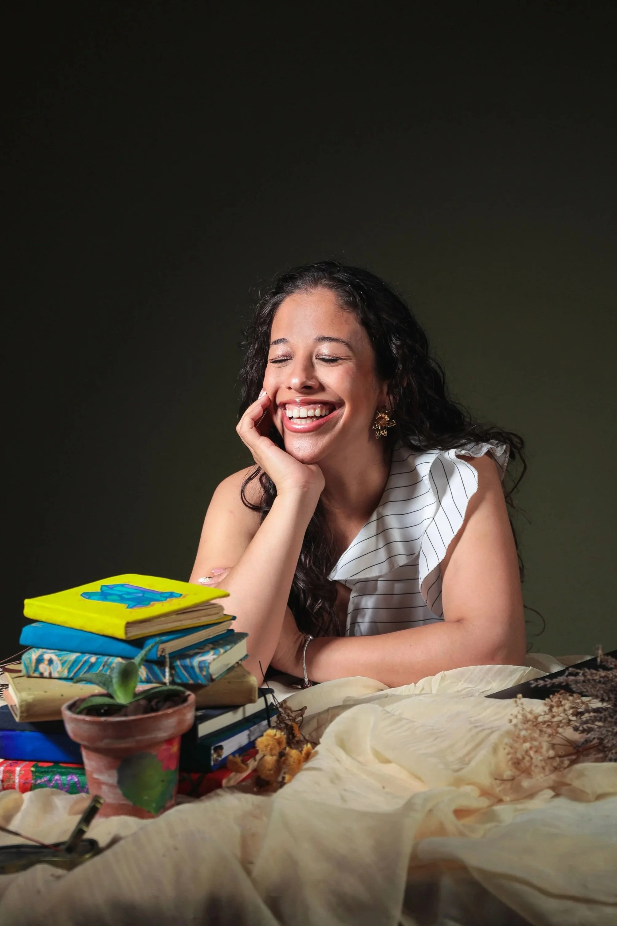 A woman with dark, curly hair and earrings is smiling with her eyes closed, resting her face on her hand. She is sitting at a table with a stack of colorful books, a potted plant, and various papers or fabrics spread out.