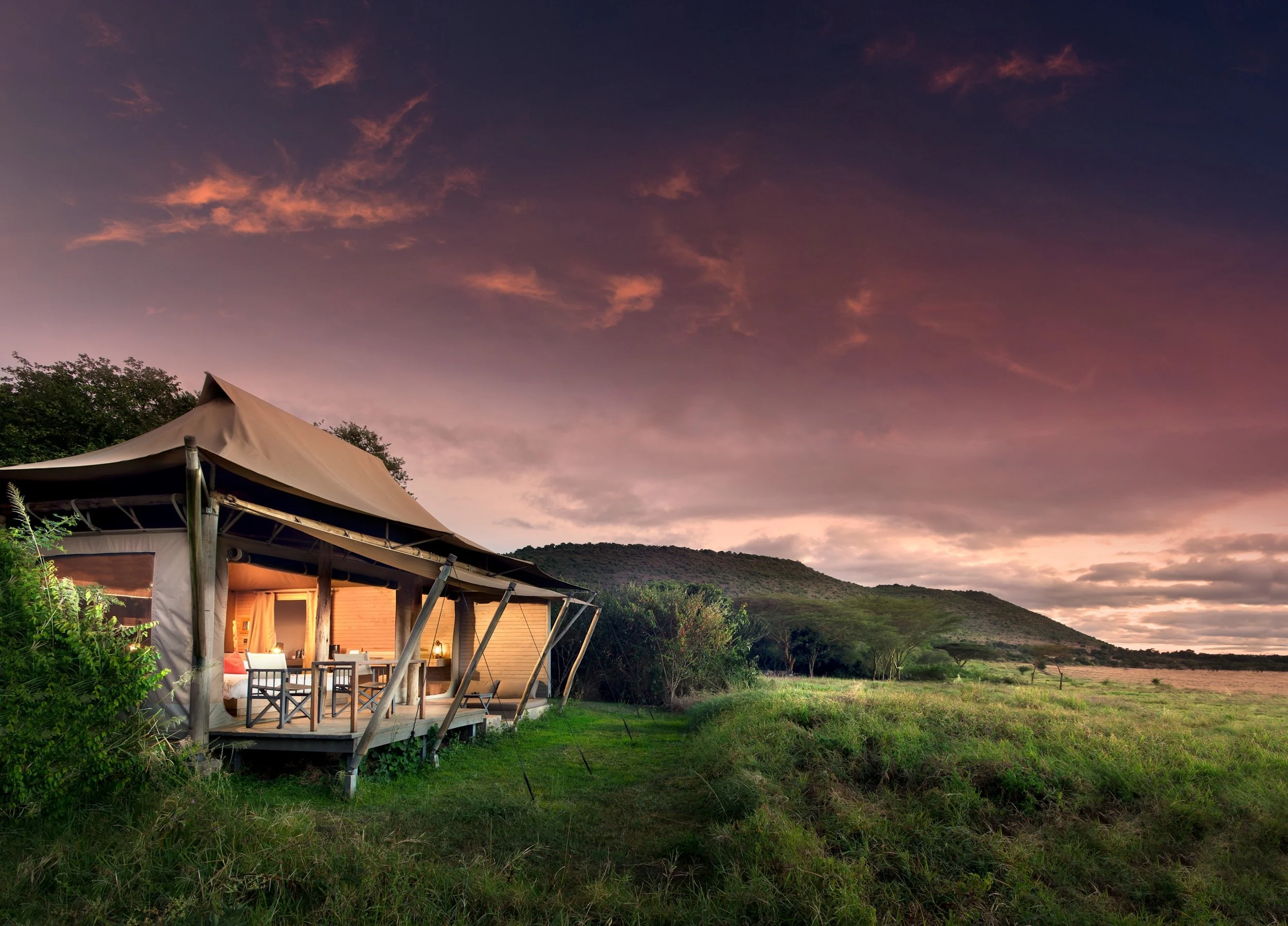 A tented lodge with an open porch, set in a grassy field with trees and distant hills in soft evening light.