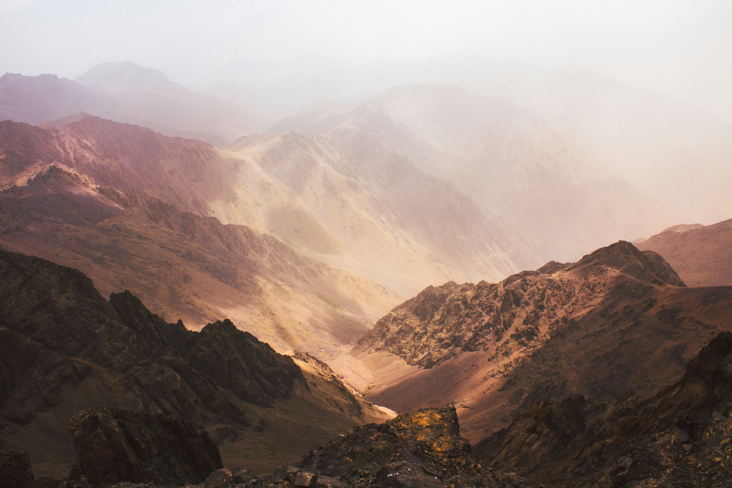 Mountain landscape with rugged peaks and ridges, some covered in light mist or clouds, in shades of brown, tan, and dark gray.