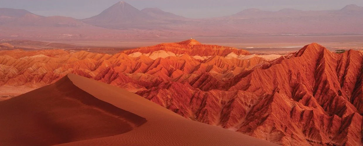 Colorful desert landscape with sand dunes and ridges, mountains in the background.