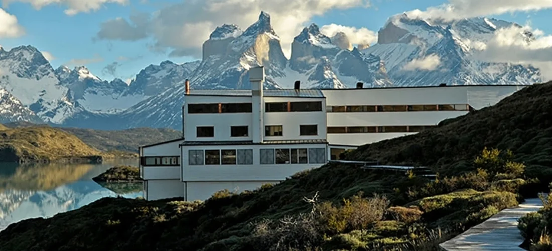 Modern white building with multiple levels, large windows, and a chimney, situated on a green hillside near a lake, with snow-capped mountains in the background.