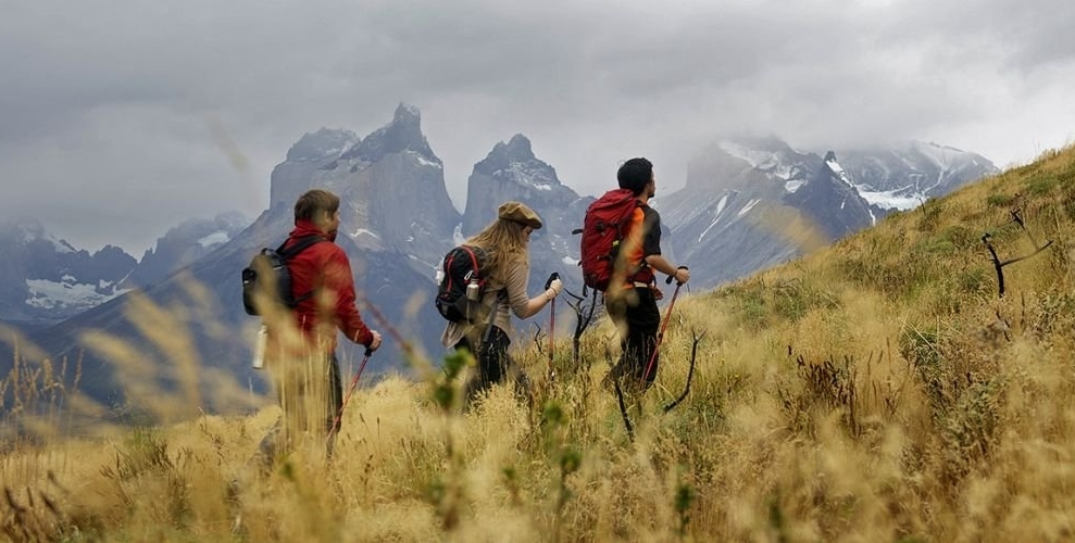 Three hikers with backpacks walking through grassy terrain with mountains in the background.