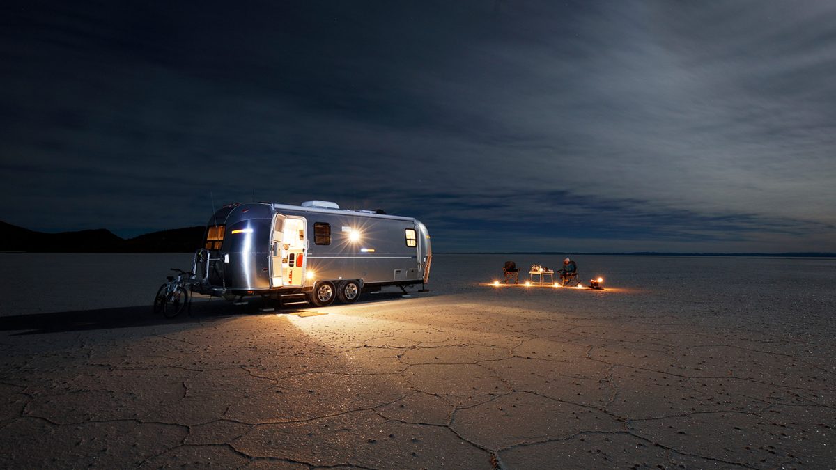 Aot camper trailer with a bike attached parked on a flat, cracked ground at dusk. Two people are sitting near a small table with candles, in a remote and open landscape under a dark, cloudy sky.