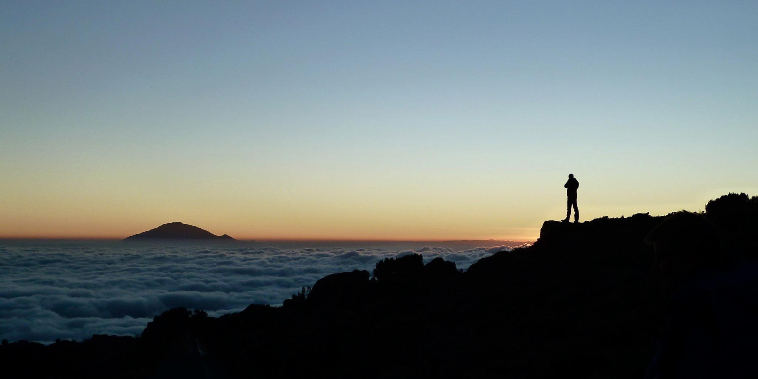 Silhouette of a person standing on a rocky ledge at sunset, overlooking a sea of clouds with a distant mountain on the horizon.