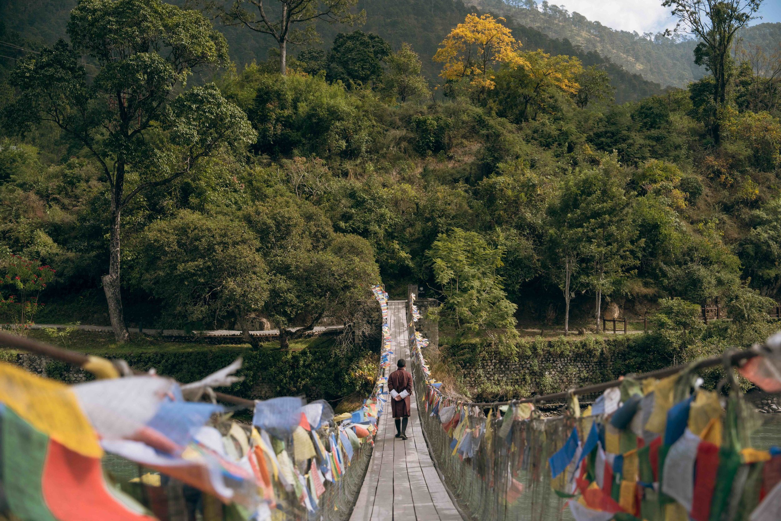 A person walking on a narrow suspension bridge decorated with colorful prayer flags, surrounded by lush green trees and mountains in the background.