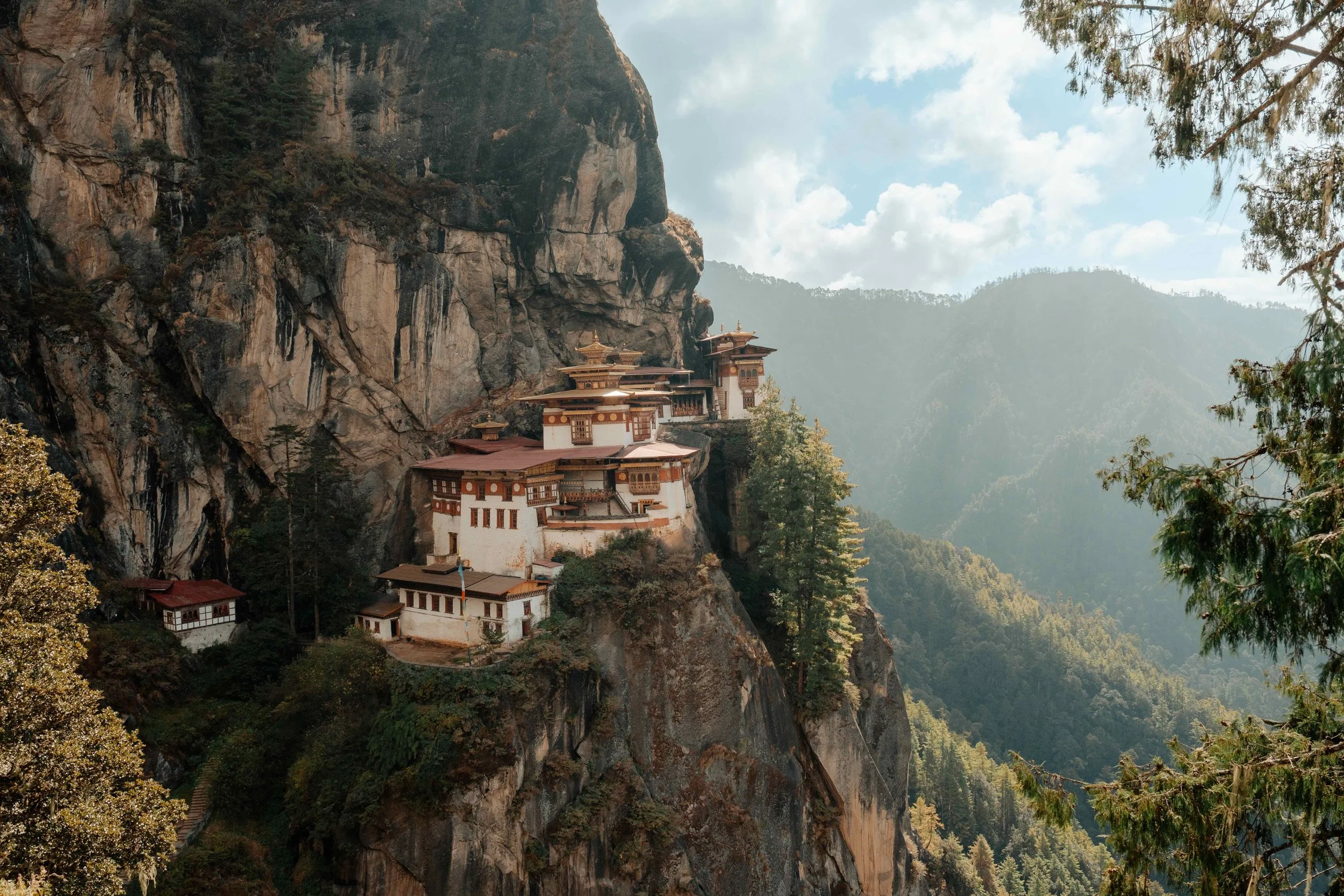 A monastery built into a cliffside, surrounded by forested mountains on a sunny day with a partly cloudy sky.