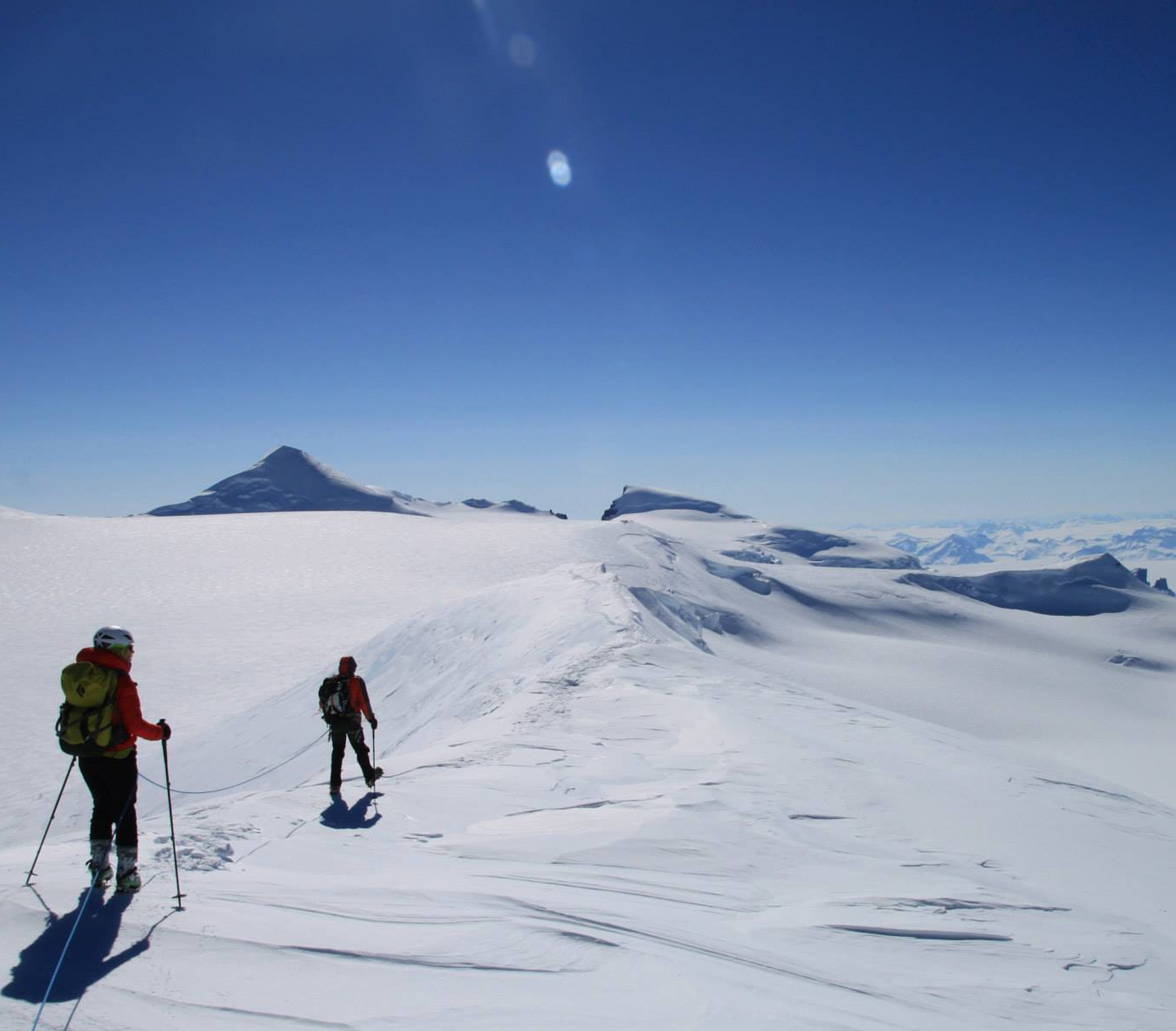 Two mountaineers in red jackets hiking across a snow-covered landscape under a clear blue sky.