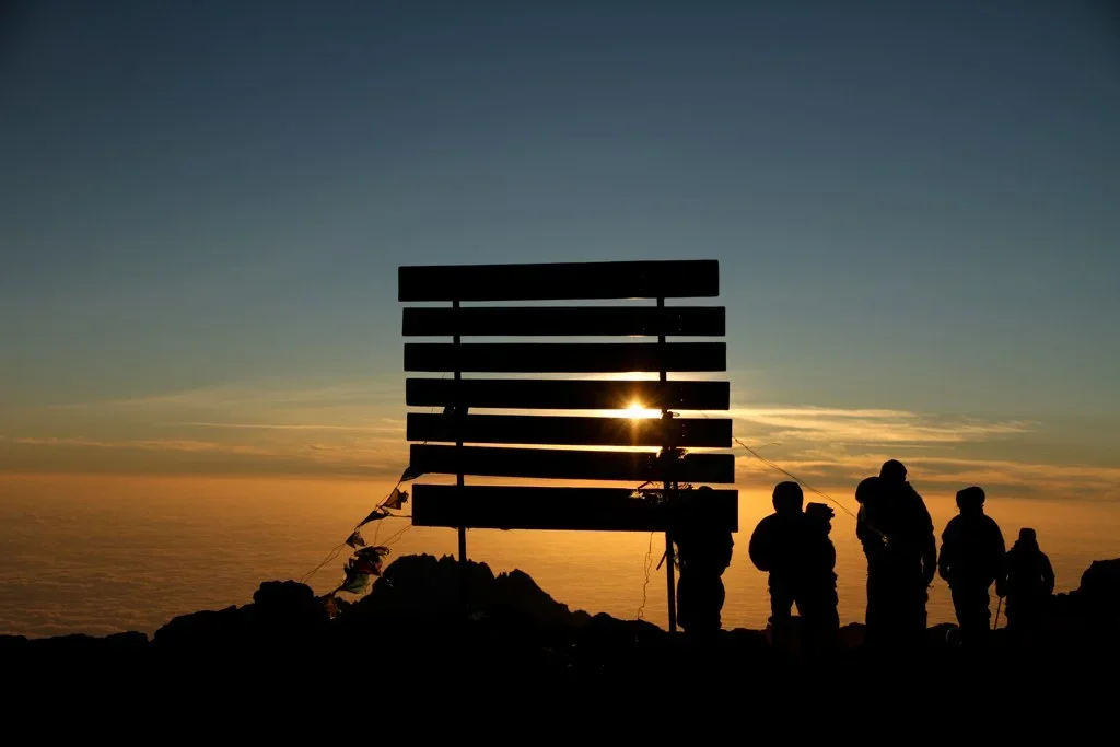People standing on a hill at sunset with a wooden structure, possibly a small shed or art installation, silhouetted against the sky and ocean.