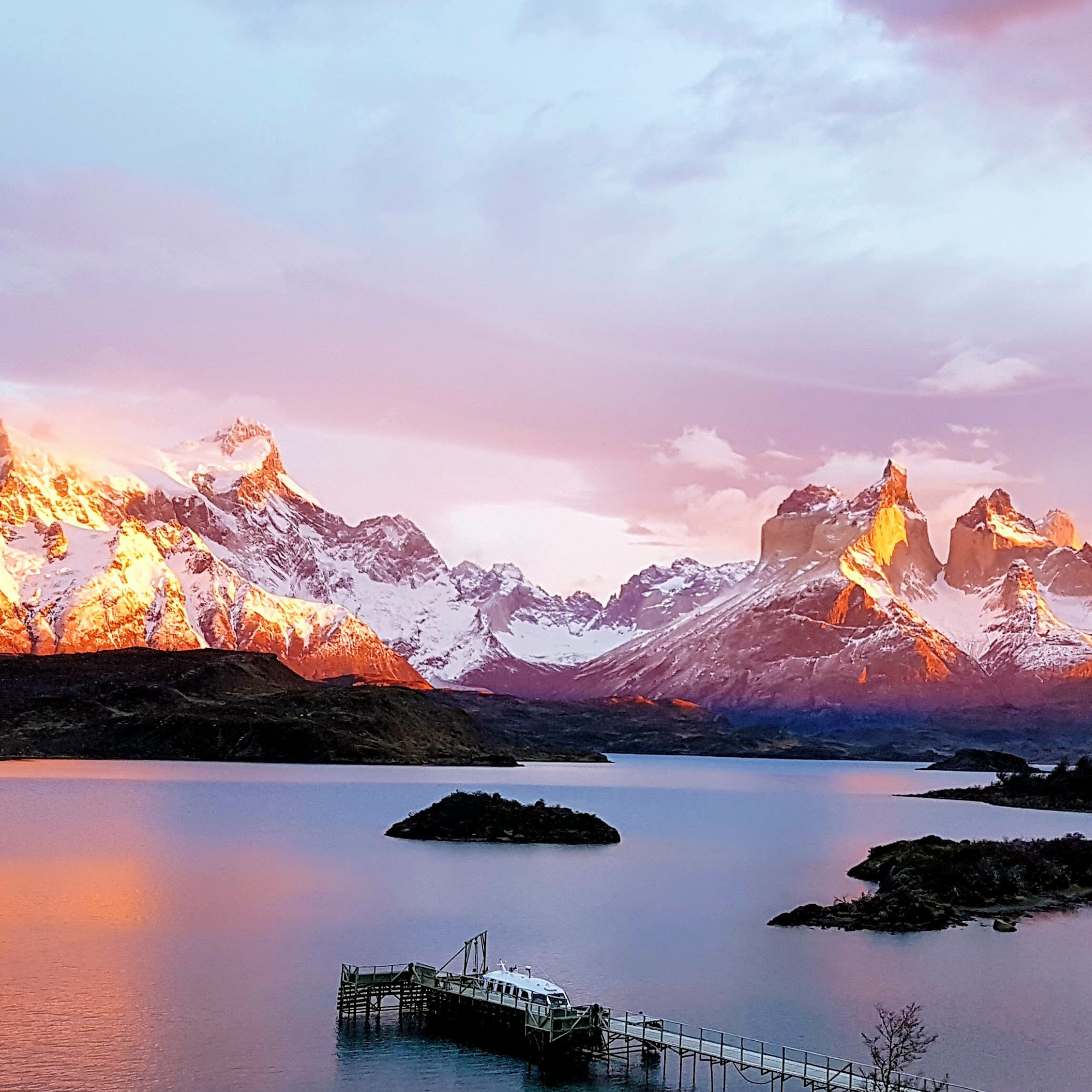 Sunset over snow-capped mountains reflecting on a calm lake with a small boat dock and islands in the foreground.