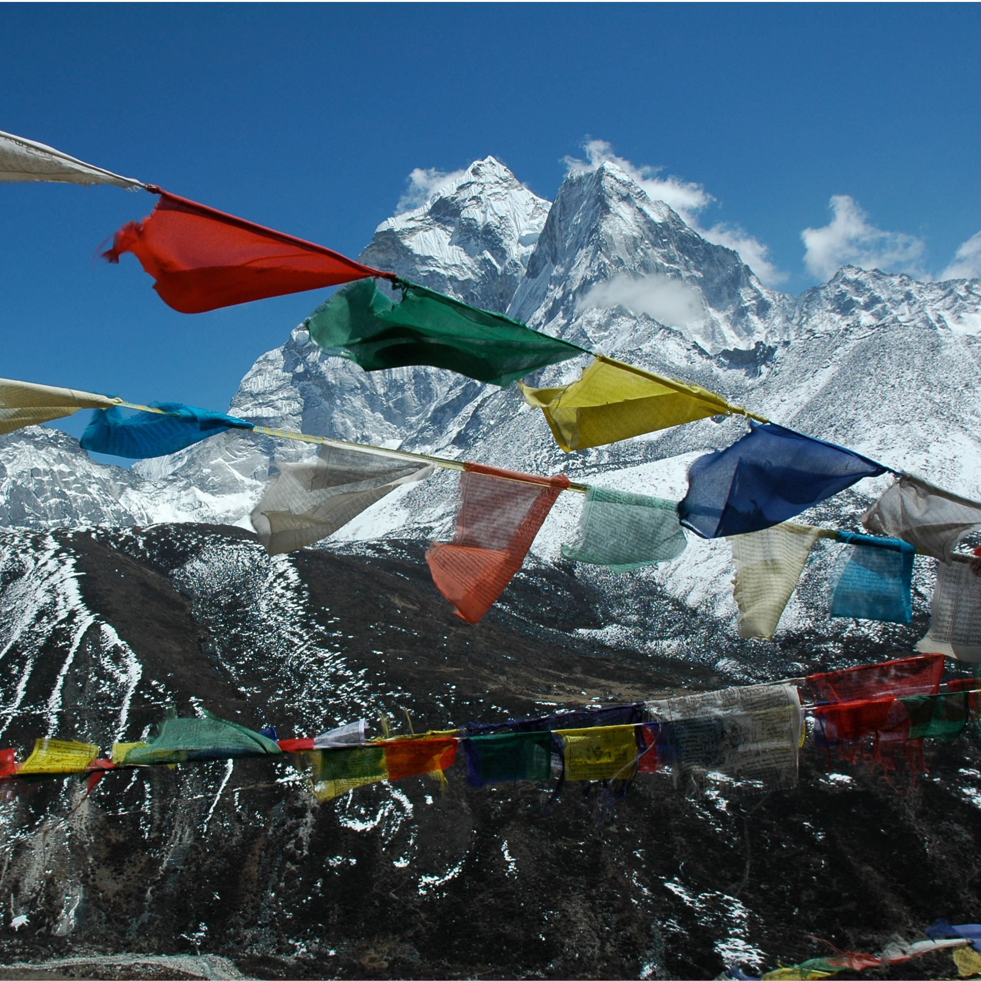 Colorful prayer flags strung across snow-capped mountain peaks under a clear blue sky, with rugged terrain below.