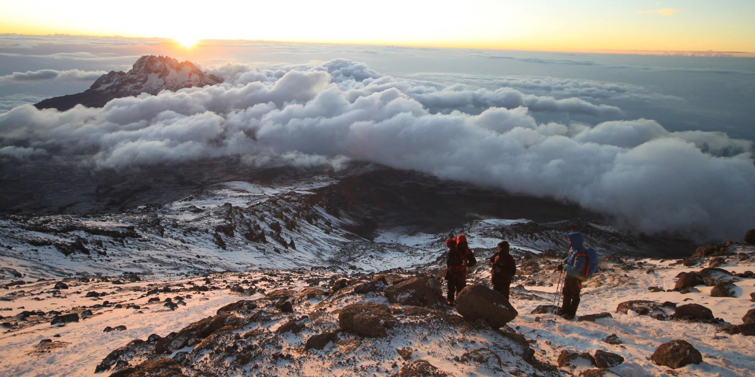 Three hikers in winter gear standing on a snowy mountain slope with clouds and a mountain in the background during sunrise.