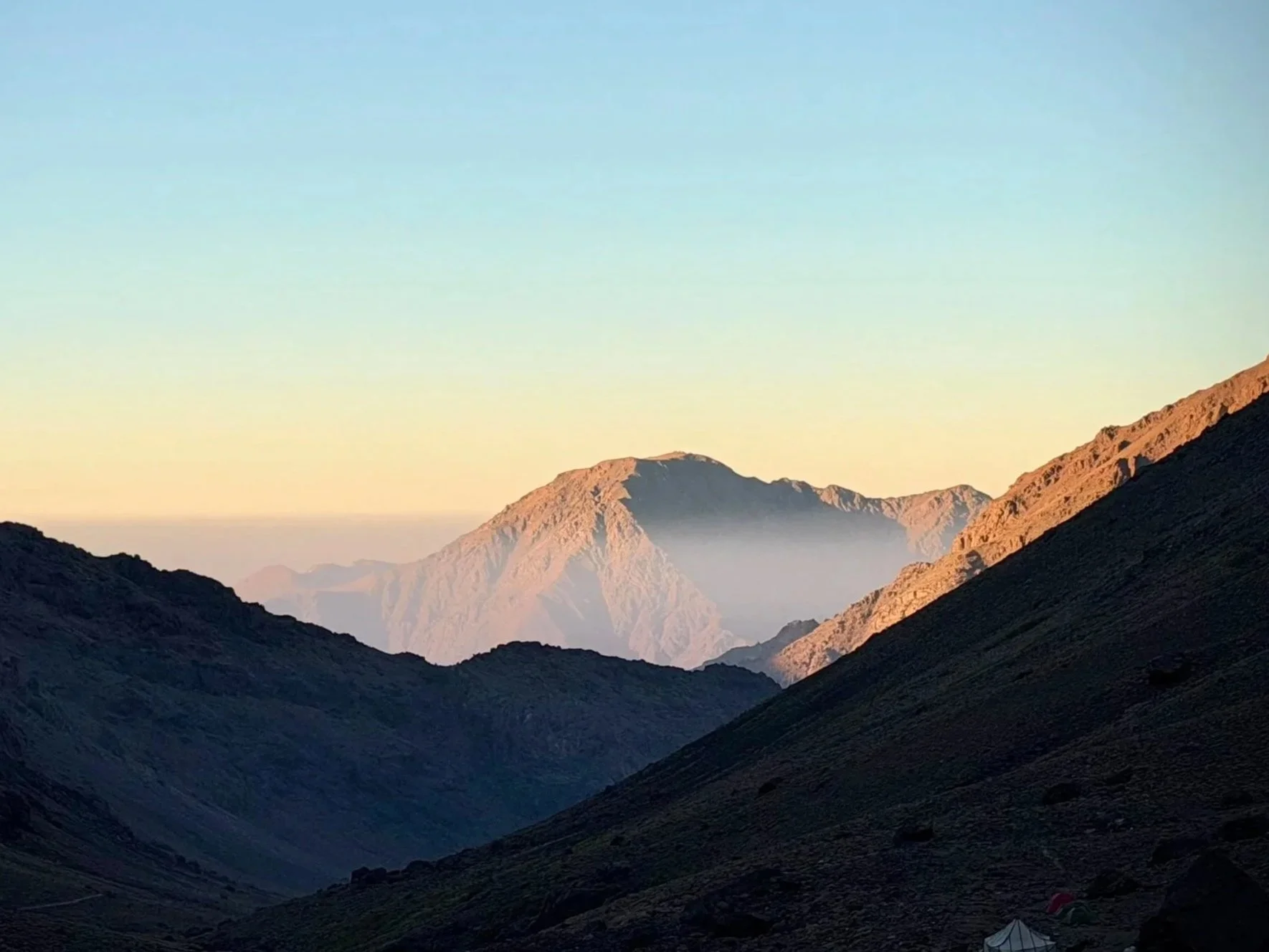 Mountain landscape at sunrise with peaks and valleys, some with snow, and a clear sky.